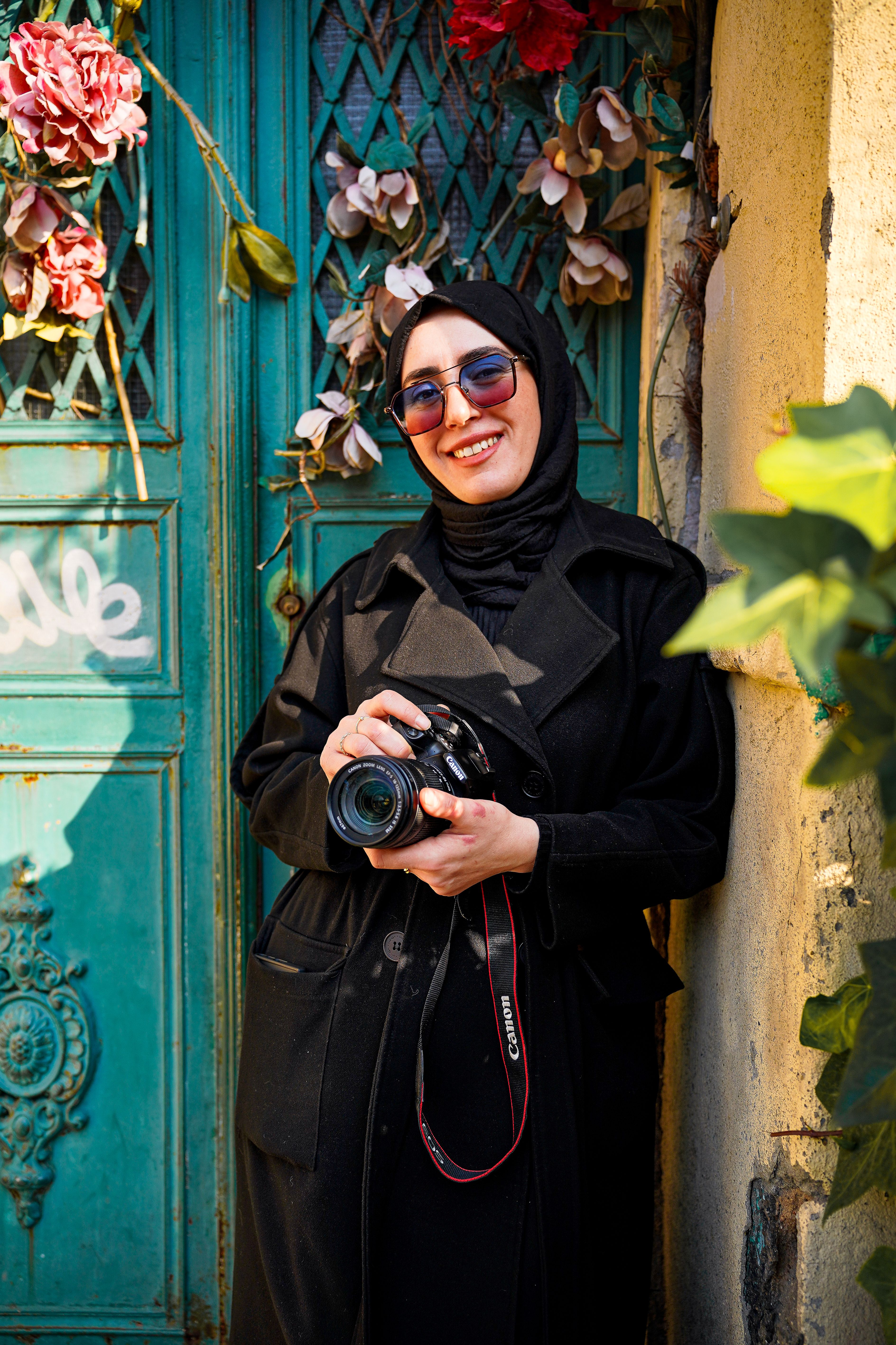 Smiling photographer holding camera in front of vintage door with floral decor.