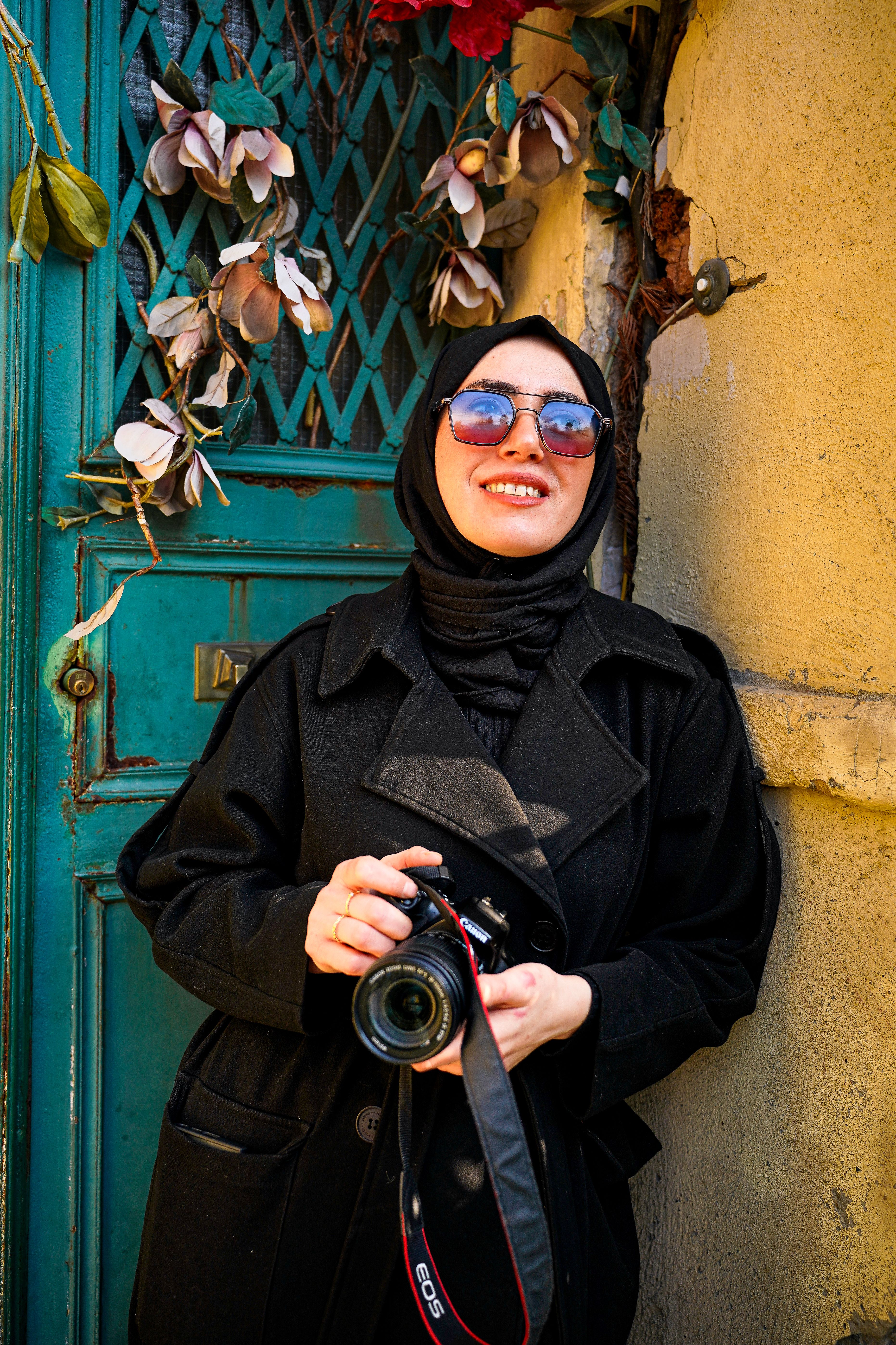 Smiling woman in hijab holding a camera against rustic outdoor background.