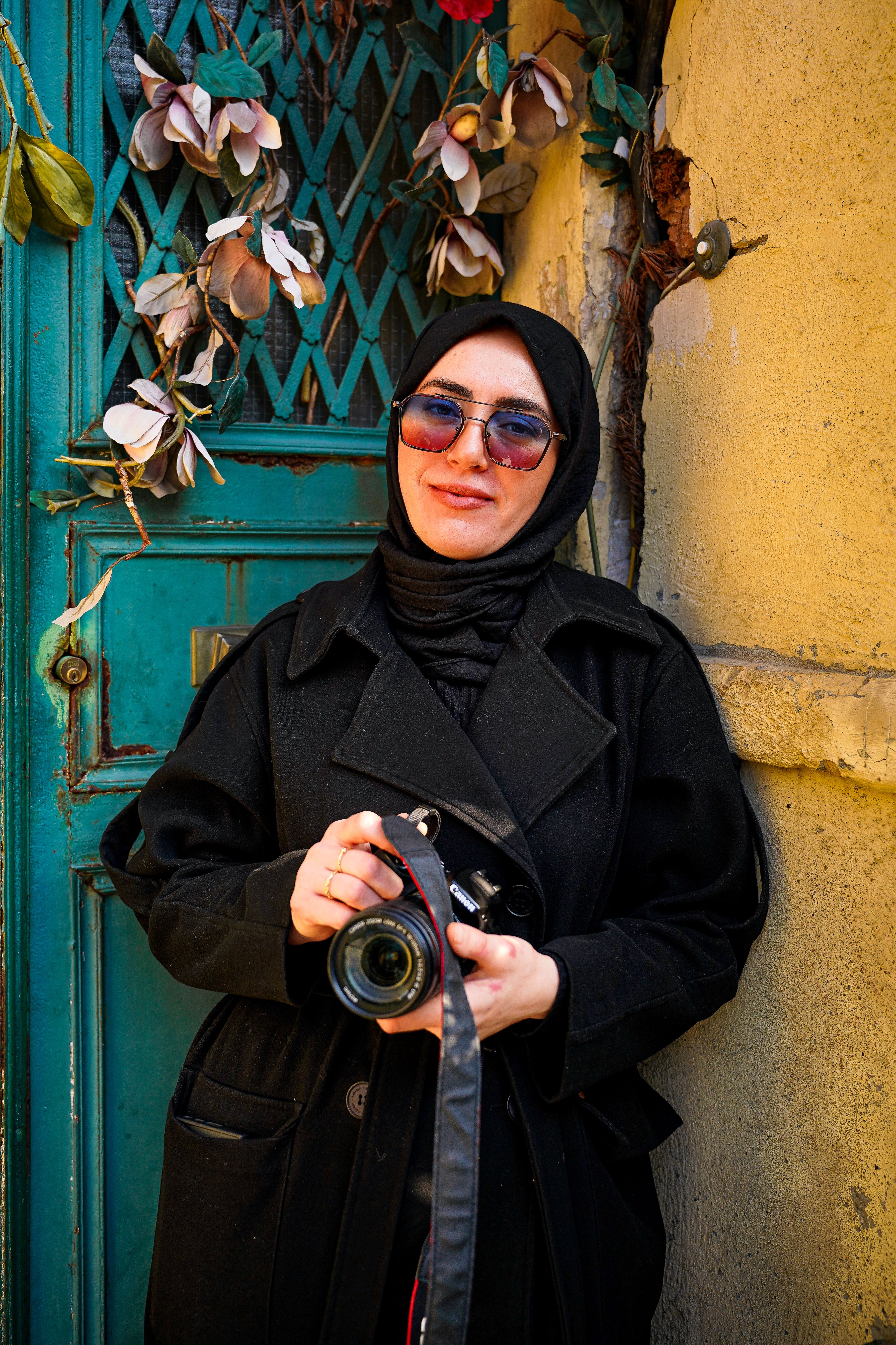 A woman wearing sunglasses and a hijab, holding a camera, stands confidently against a rustic gate.