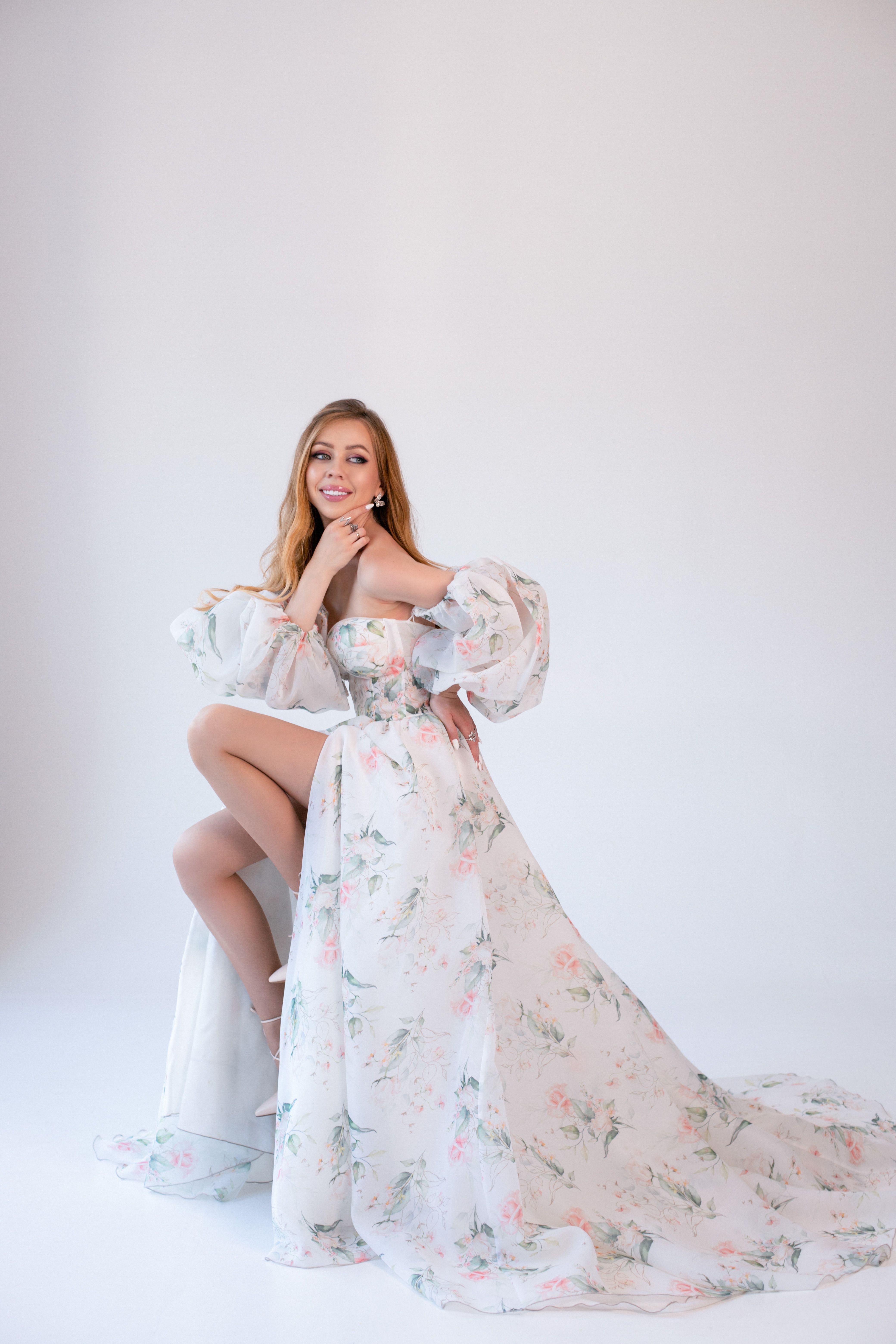 A stylish woman poses gracefully in a white floral gown with puff sleeves, exuding elegance.