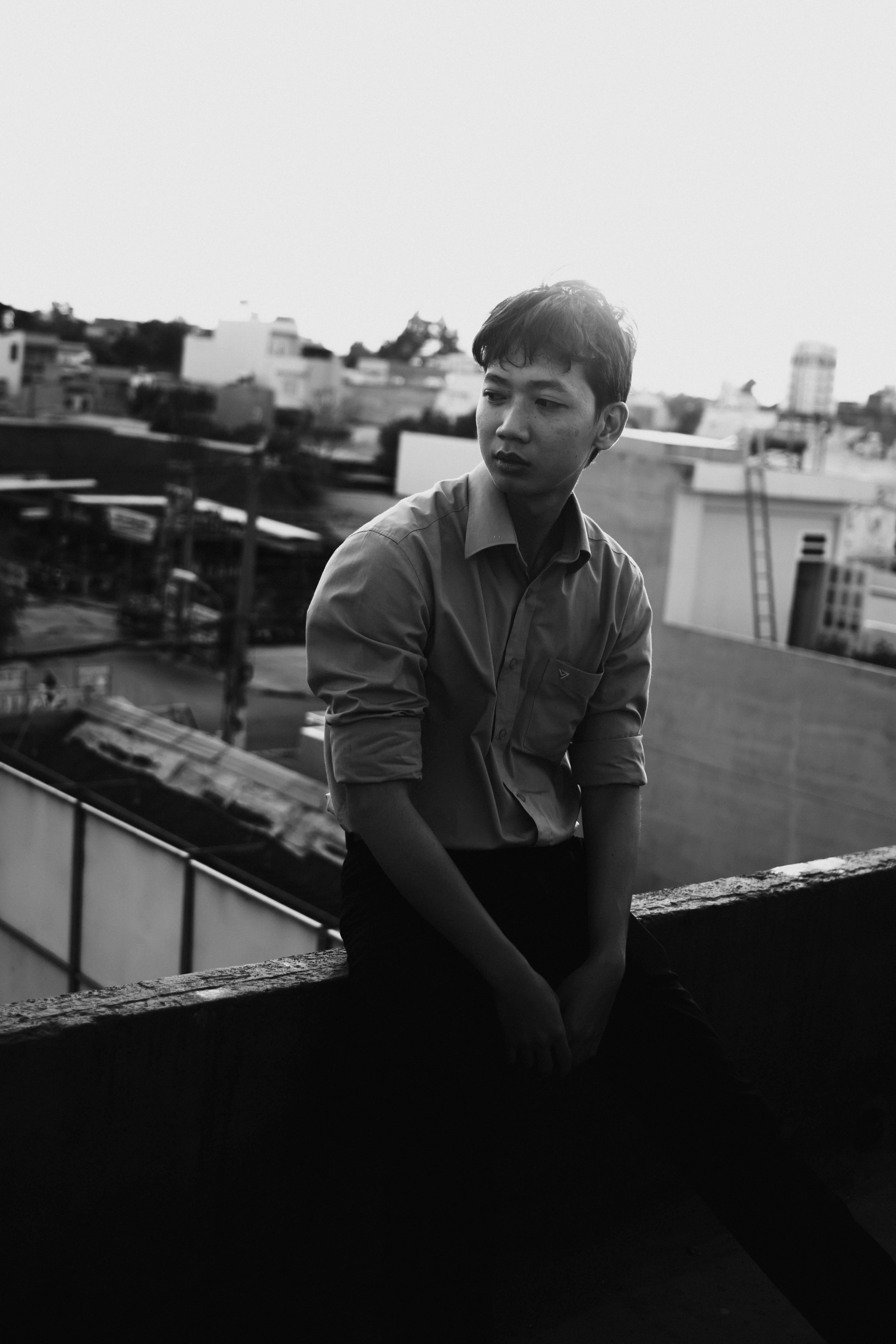 A black and white portrait of a man sitting on a rooftop ledge in an urban setting.