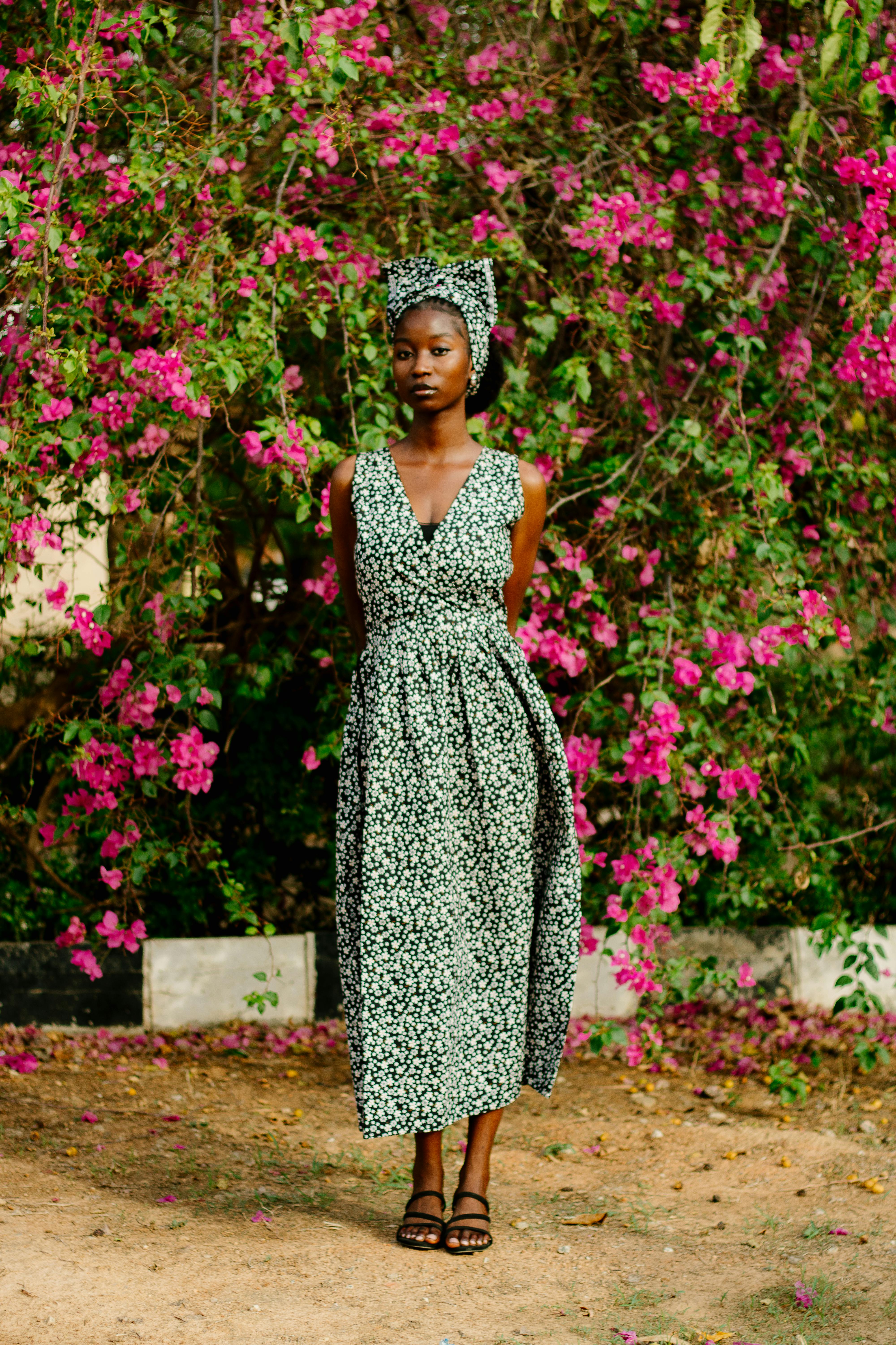 A woman in a floral dress stands against a backdrop of vivid pink flowers outdoors.