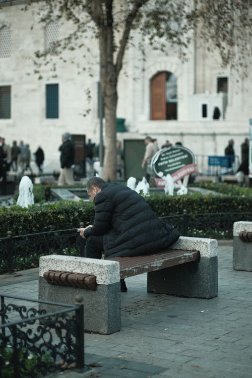 Free A solitary man in a black coat sits on a park bench in an urban setting, exuding a serene and relaxed atmosphere. Stock Photo