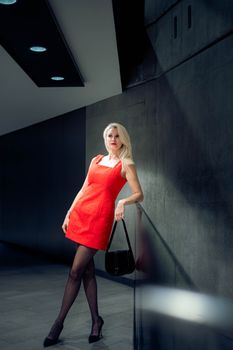 Elegant woman in red dress poses stylishly against a dark wall, holding a purse.