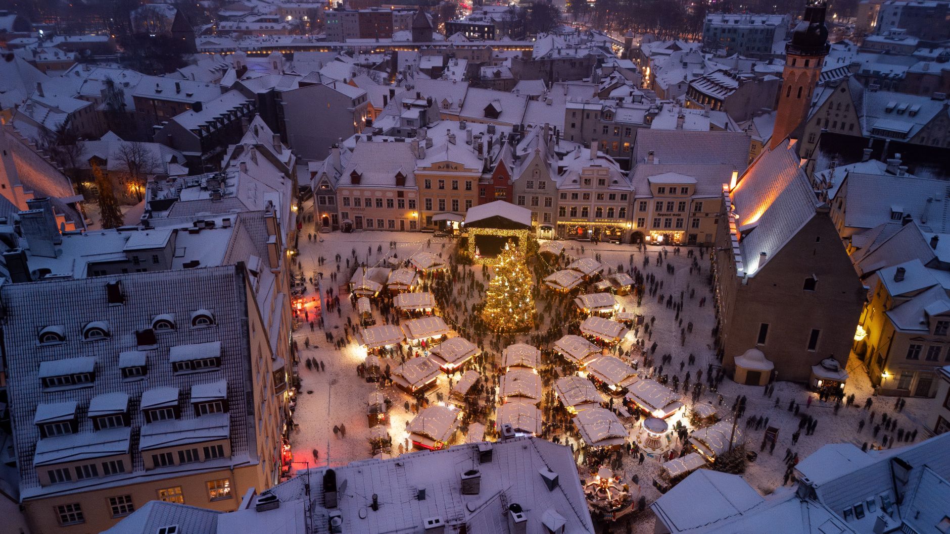 Tallinn's Christmas market on Town Hall Square at dusk, lit stalls and snow-covered roofs seen from above