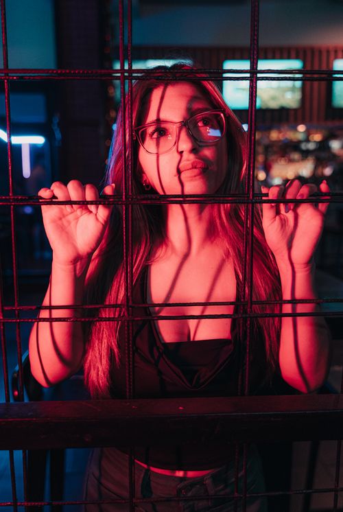 Free Portrait of a young woman in eyeglasses behind bars with vibrant red lighting at night in a club. Stock Photo