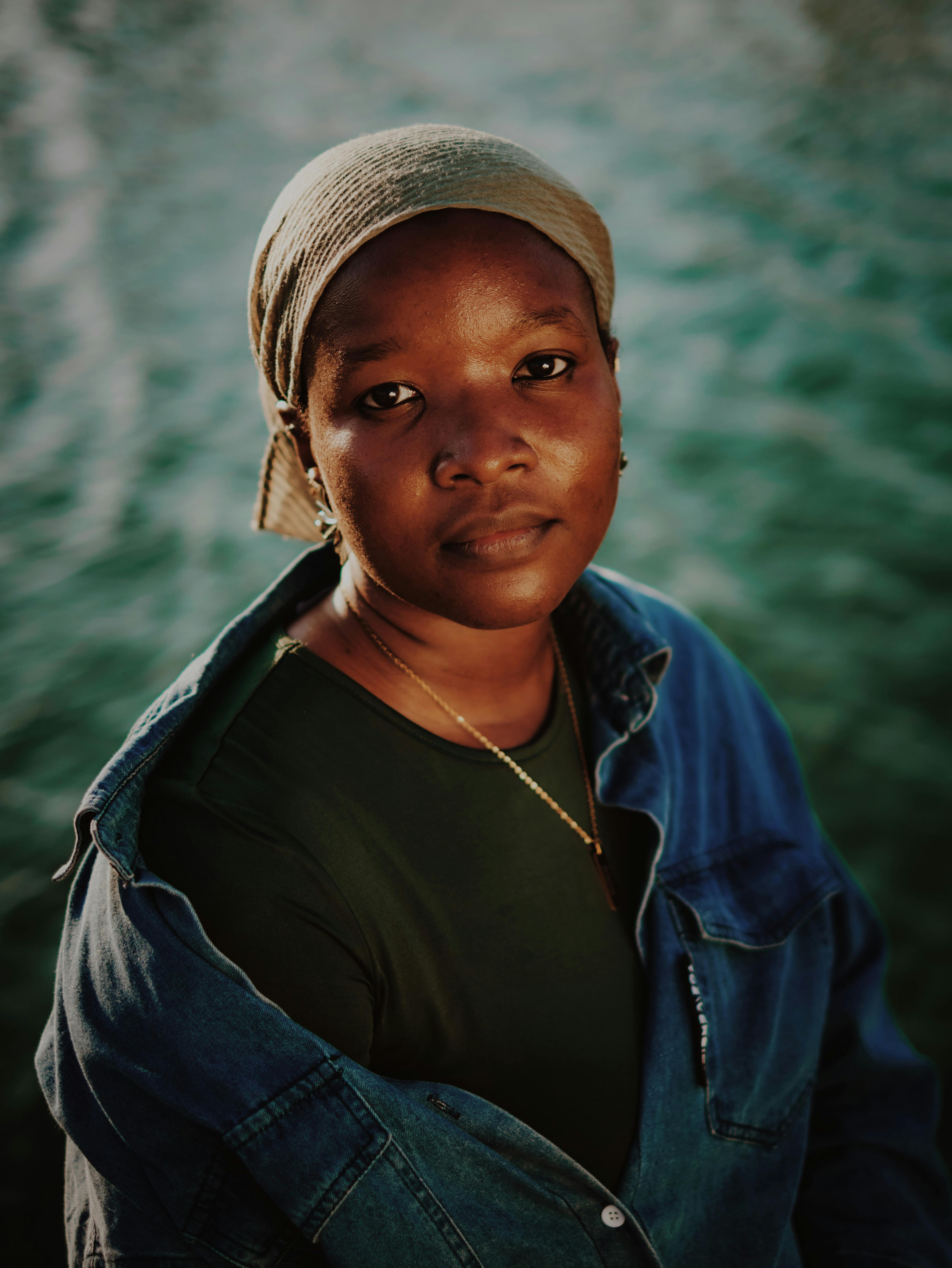 Thoughtful woman in denim jacket and headscarf by the water at sunset.