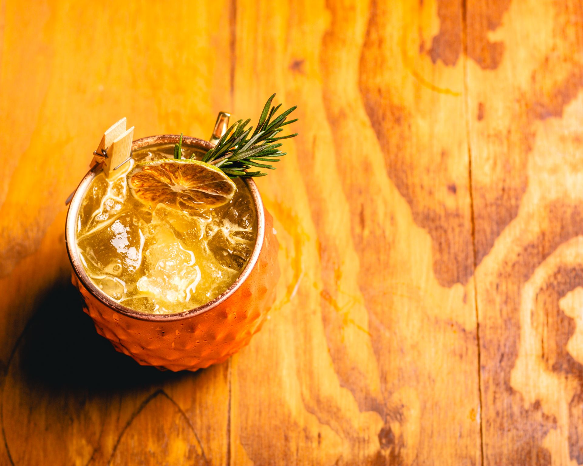 Copper mug with cocktail garnished with citrus slice and rosemary sprig on wooden table.