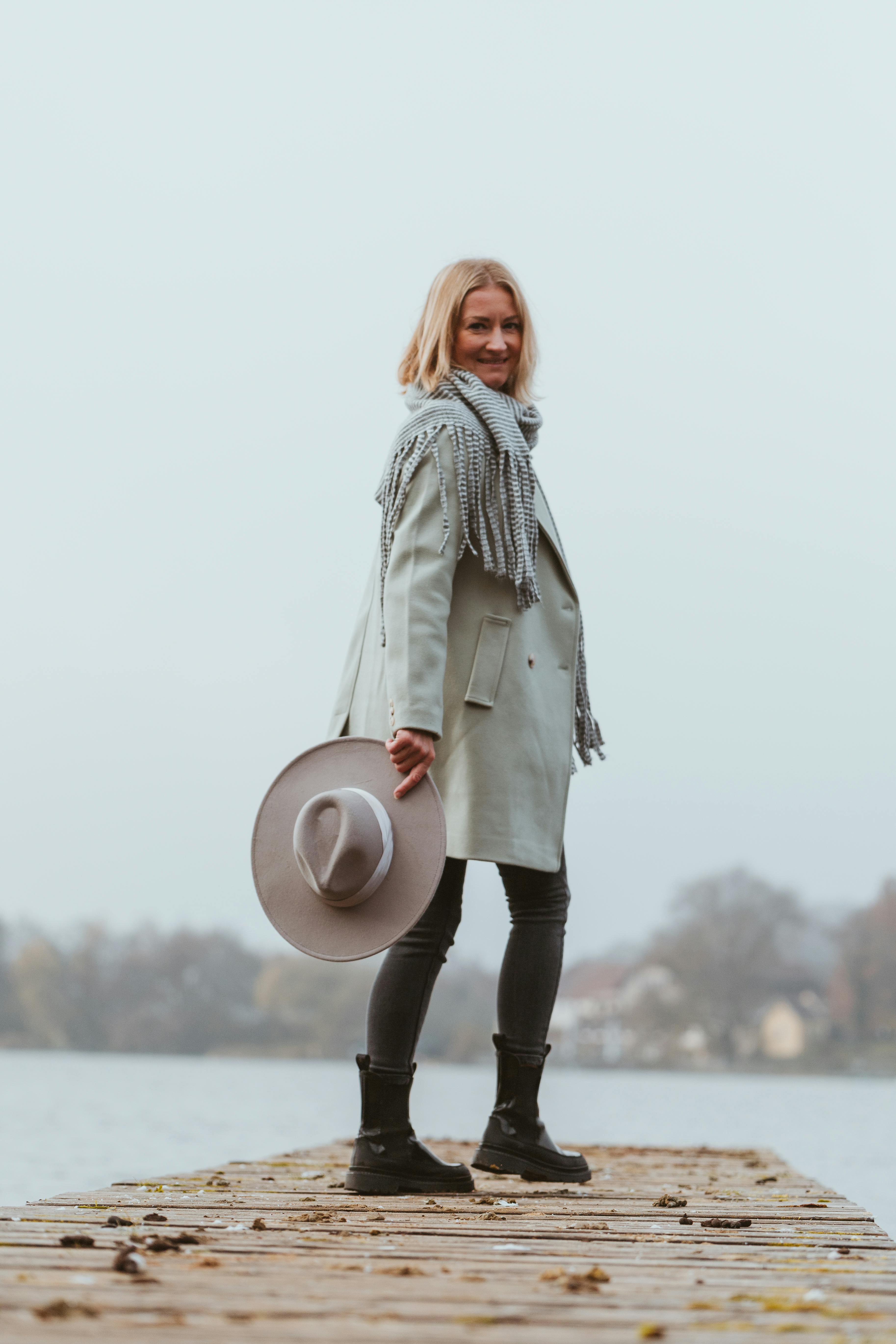 stylish woman holding hat on lakeside pier