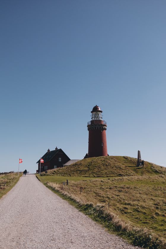A red brick lighthouse above a gravel path climbing a grassy headland — photo by Margarita on Pexels