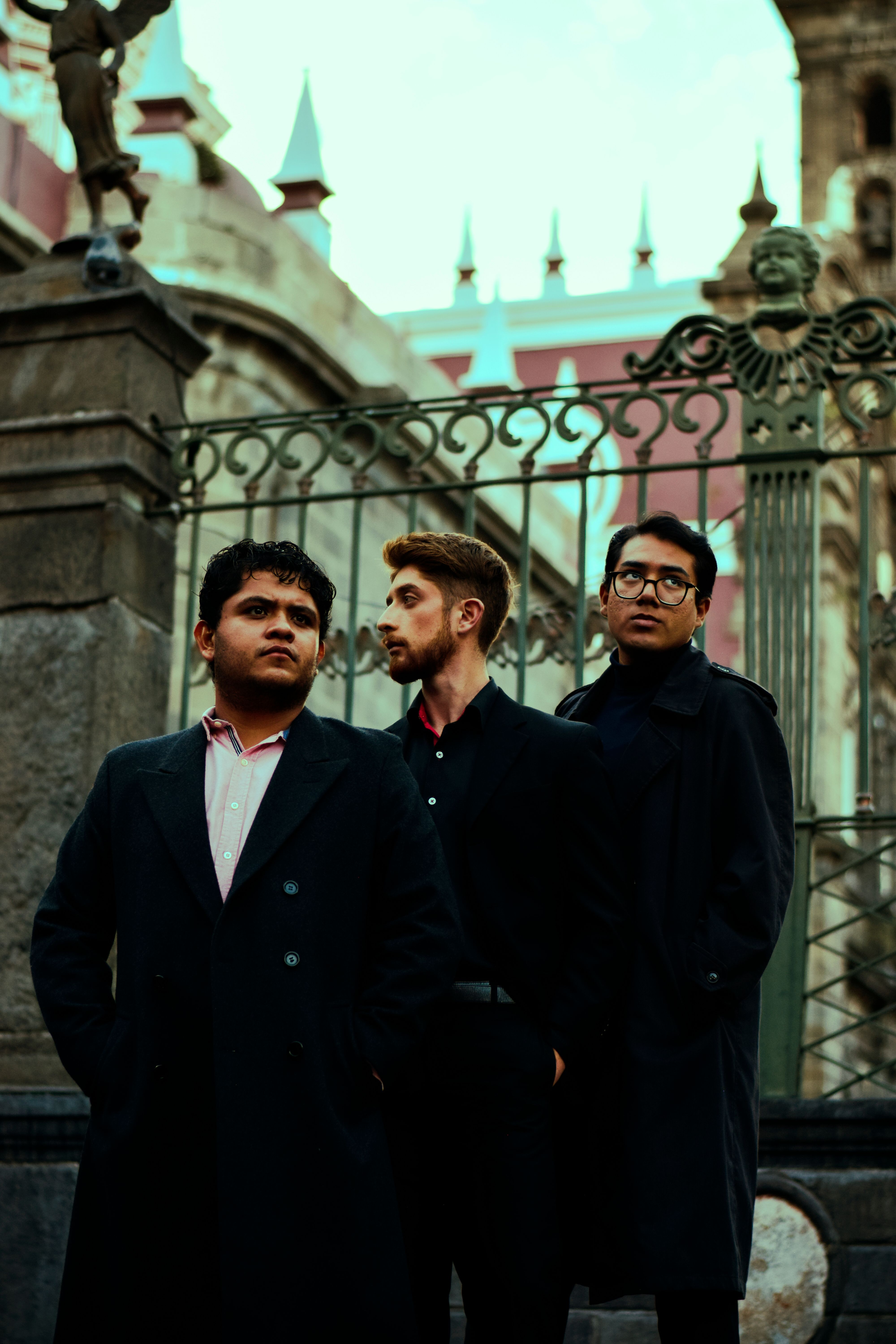 Three men in dark coats standing confidently near a decorative iron gate.