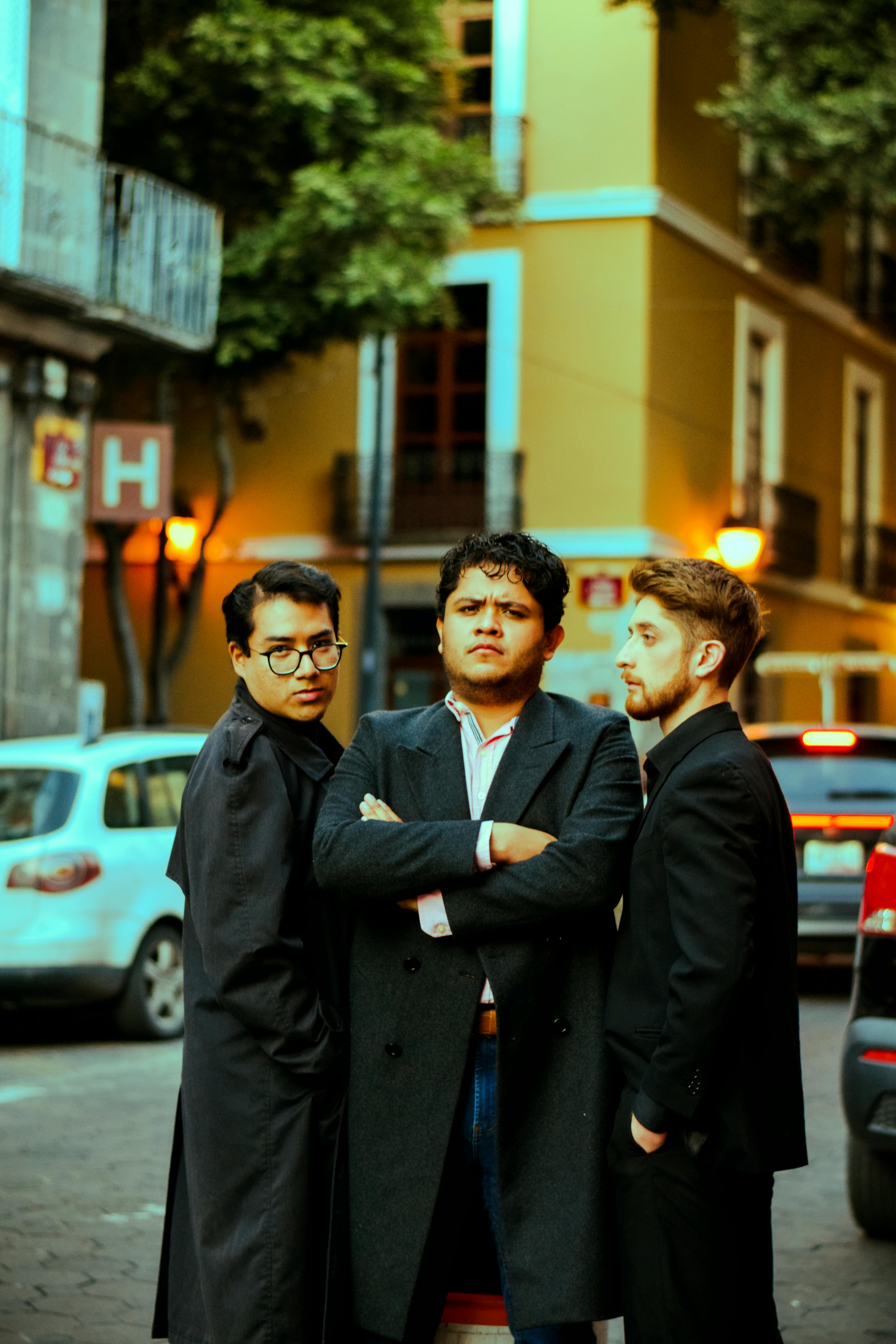 Three men pose confidently in a lively street at dusk with urban lights in the background.