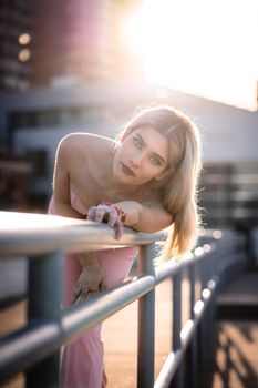 Elegant woman in pink dress leaning on rail in an urban setting with sunset backlight, capturing city fashion vibe.