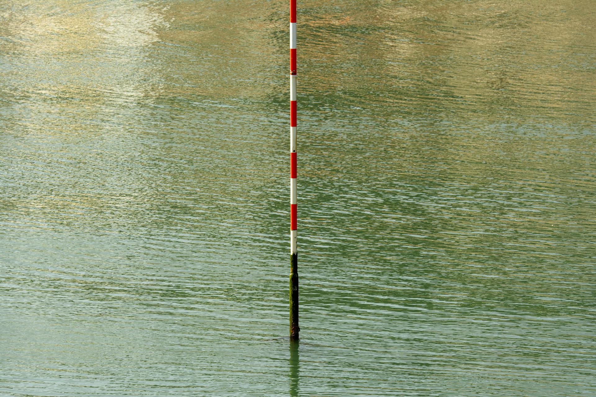A red and white post stands in calm water, reflecting stillness and measurement.