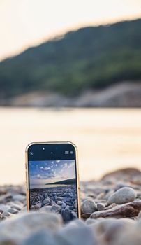A smartphone capturing a scenic view of a pebble beach with blurred hills in the background.