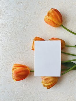 Flat lay of a blank card surrounded by orange tulips on textured beige surface, perfect for invitations or minimalist designs.