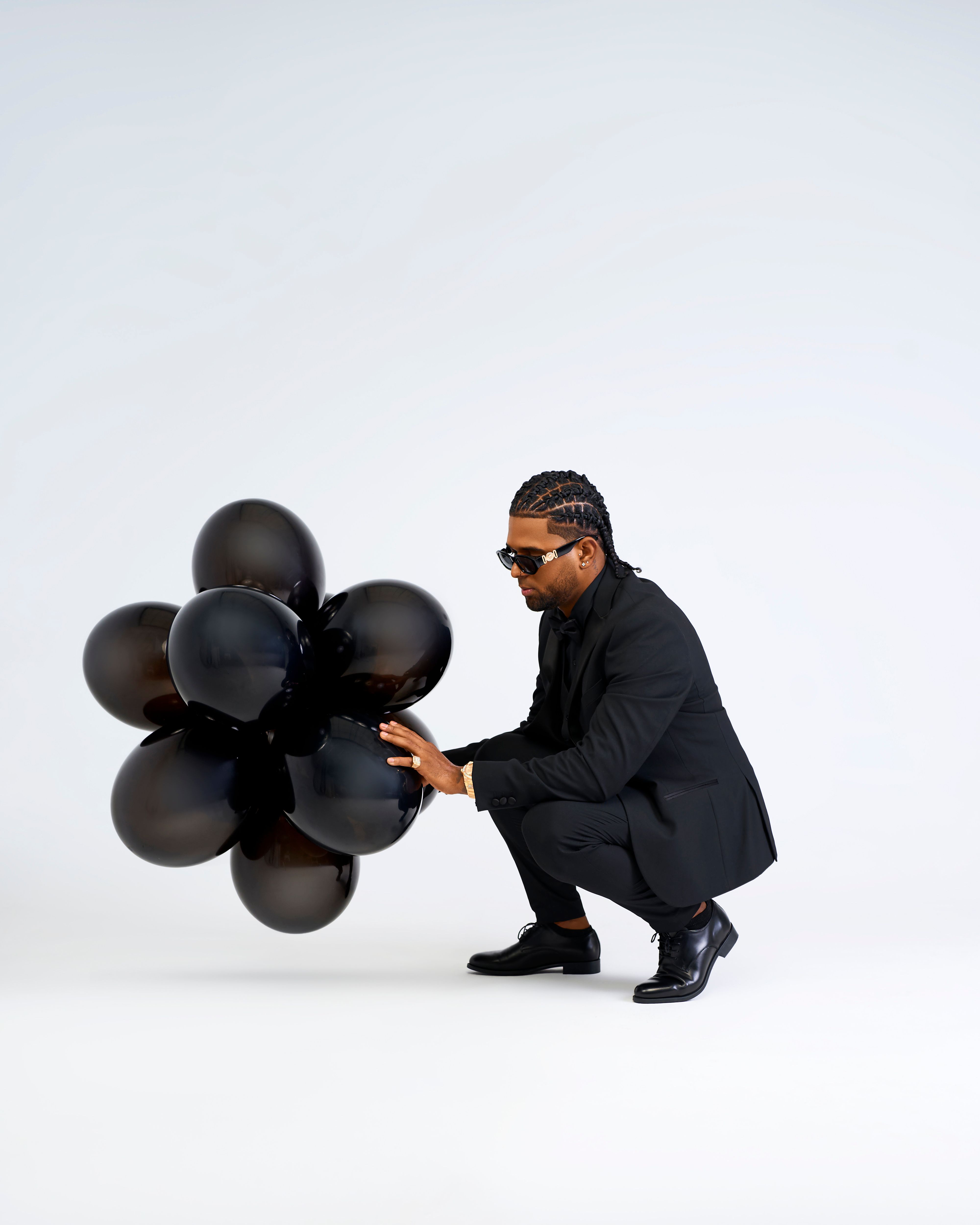 Stylish man in black kneeling with a bouquet of black balloons against a white studio background.
