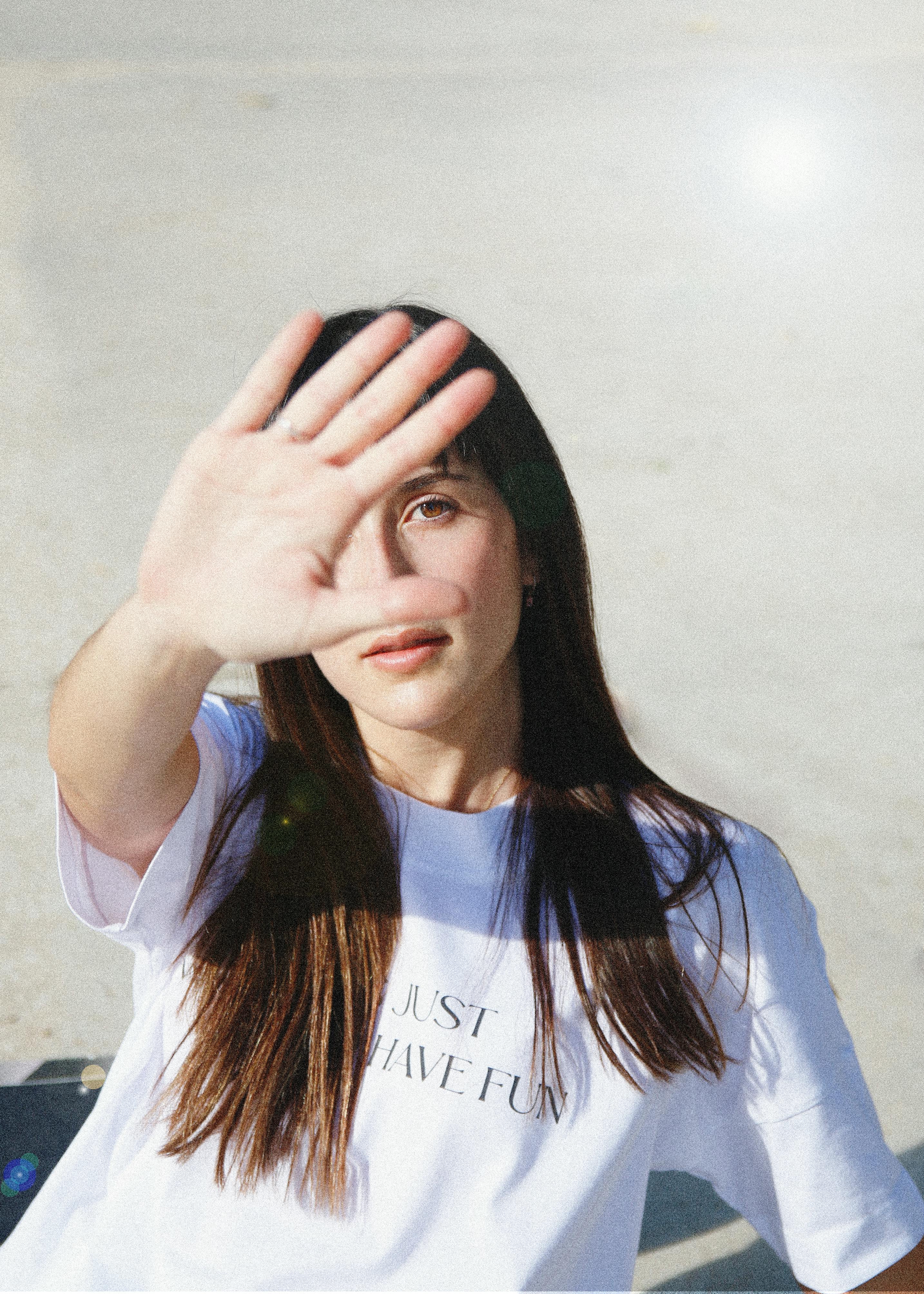 A young woman blocks the sunlight with her hand, wearing a 'Just Have Fun' t-shirt.