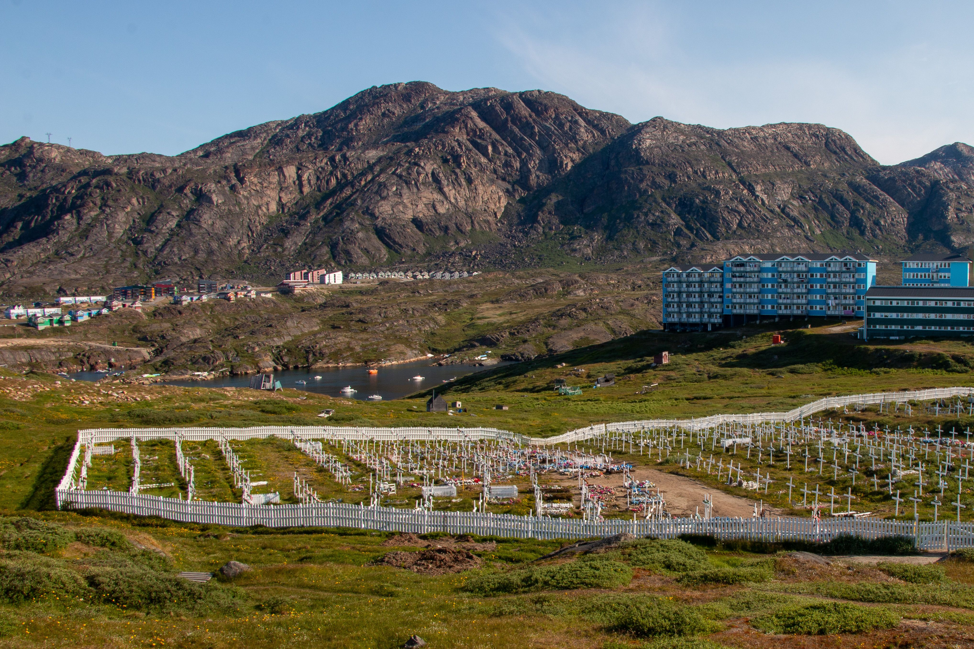 A cemetery in Sisimiut, Greenland, set against rugged mountains and modern buildings, captured on a sunny day.