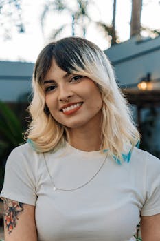 A woman with blonde hair and tattoos smiles outdoors in a peaceful garden setting.