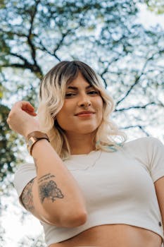 Portrait of a young woman with tattoos, blonde hair, and nose ring smiling outdoors under trees.