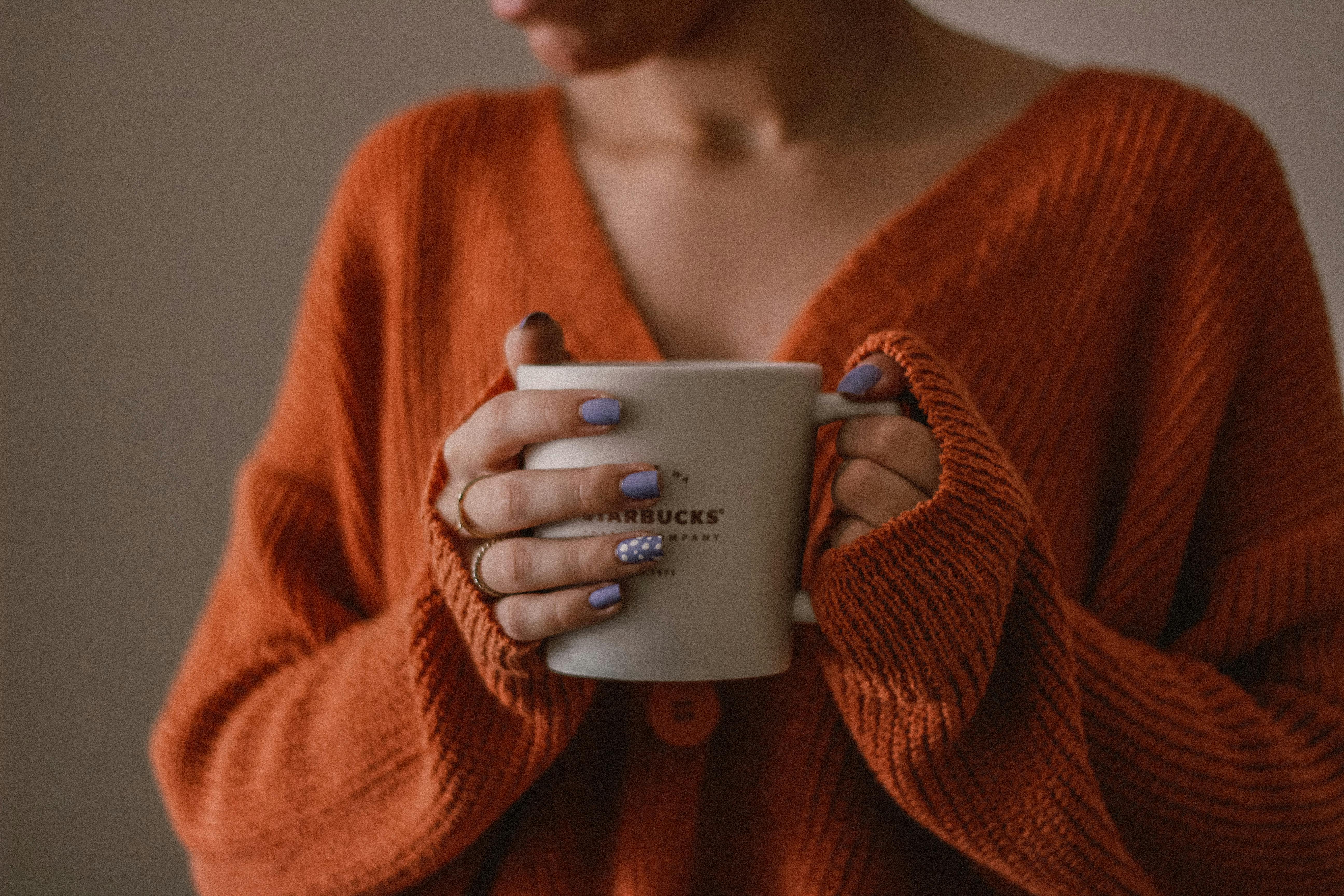Woman in orange sweater holding a warm coffee cup indoors on a cozy fall morning.