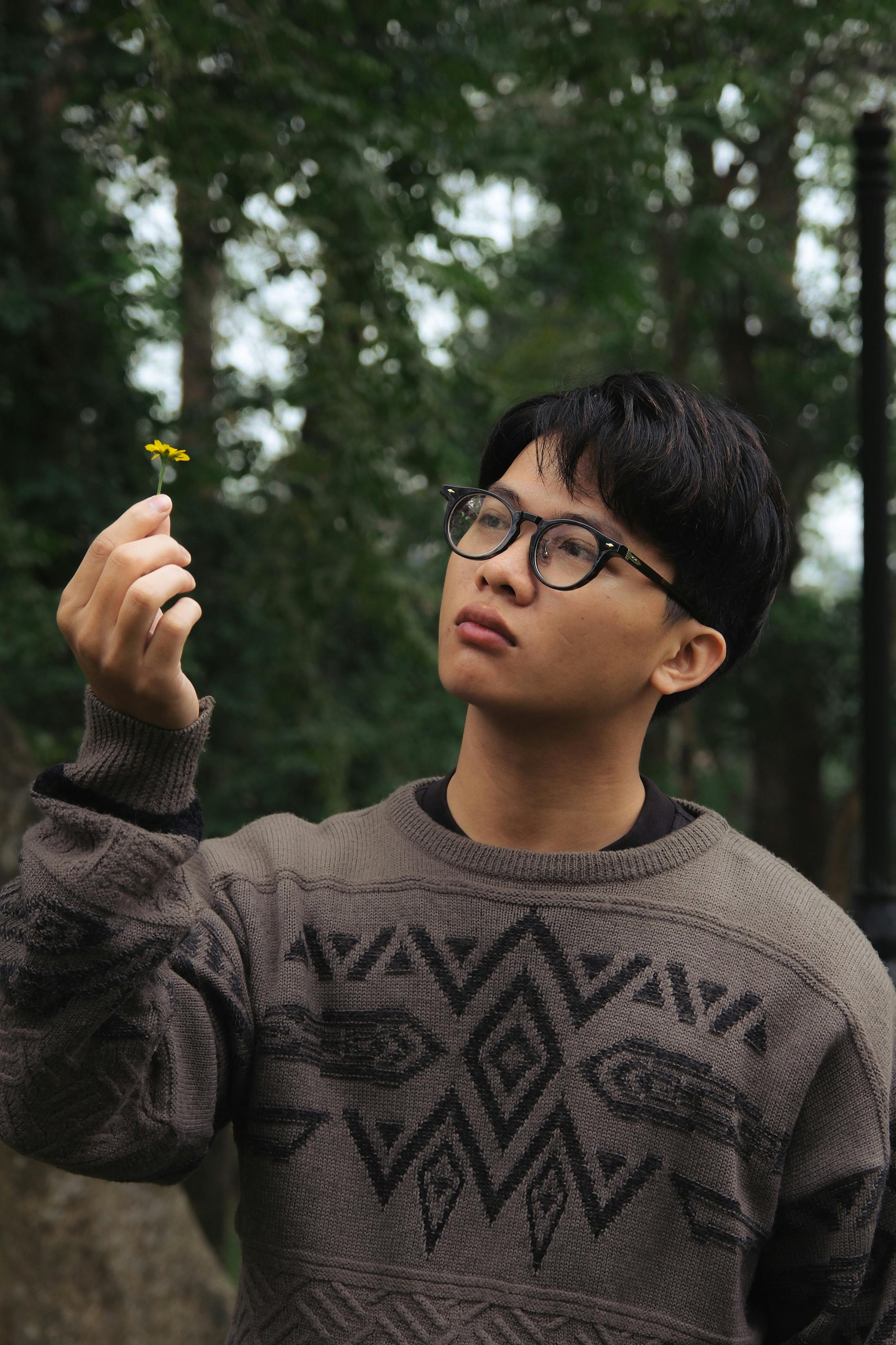 Young man wearing a patterned sweater holding a yellow flower outdoors.