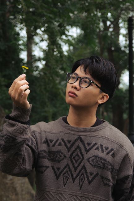 Young man wearing a patterned sweater holding a yellow flower outdoors.
