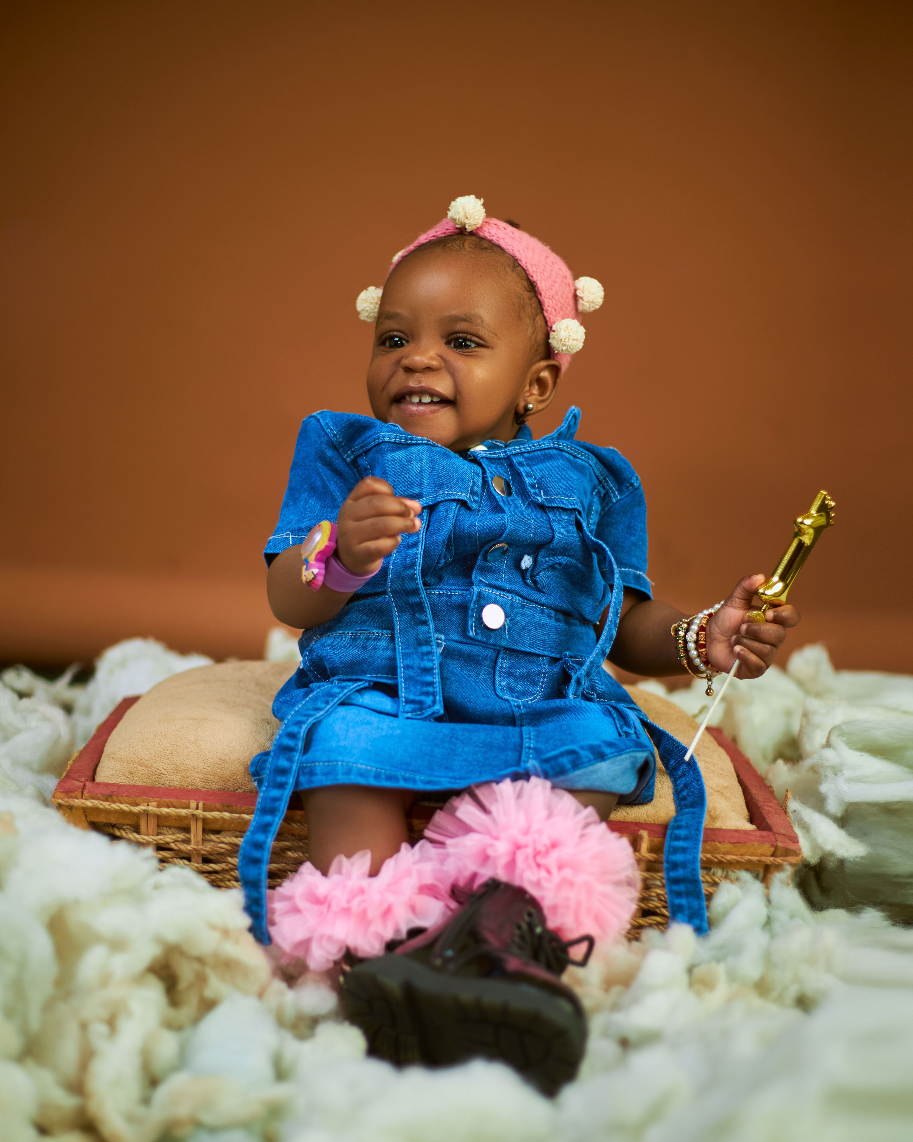 Linda menina bebê em denim, sorrindo e segurando uma varinha de brinquedo, sentada em uma almofada.
