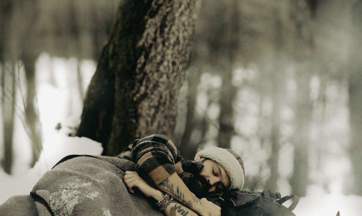 Camper resting under a thick insulating blanket beside a sleeping bag and pack in a snow-covered winter forest, illustrating how sleeping bag temperature ratings relate to overnight warmth in cold conditions