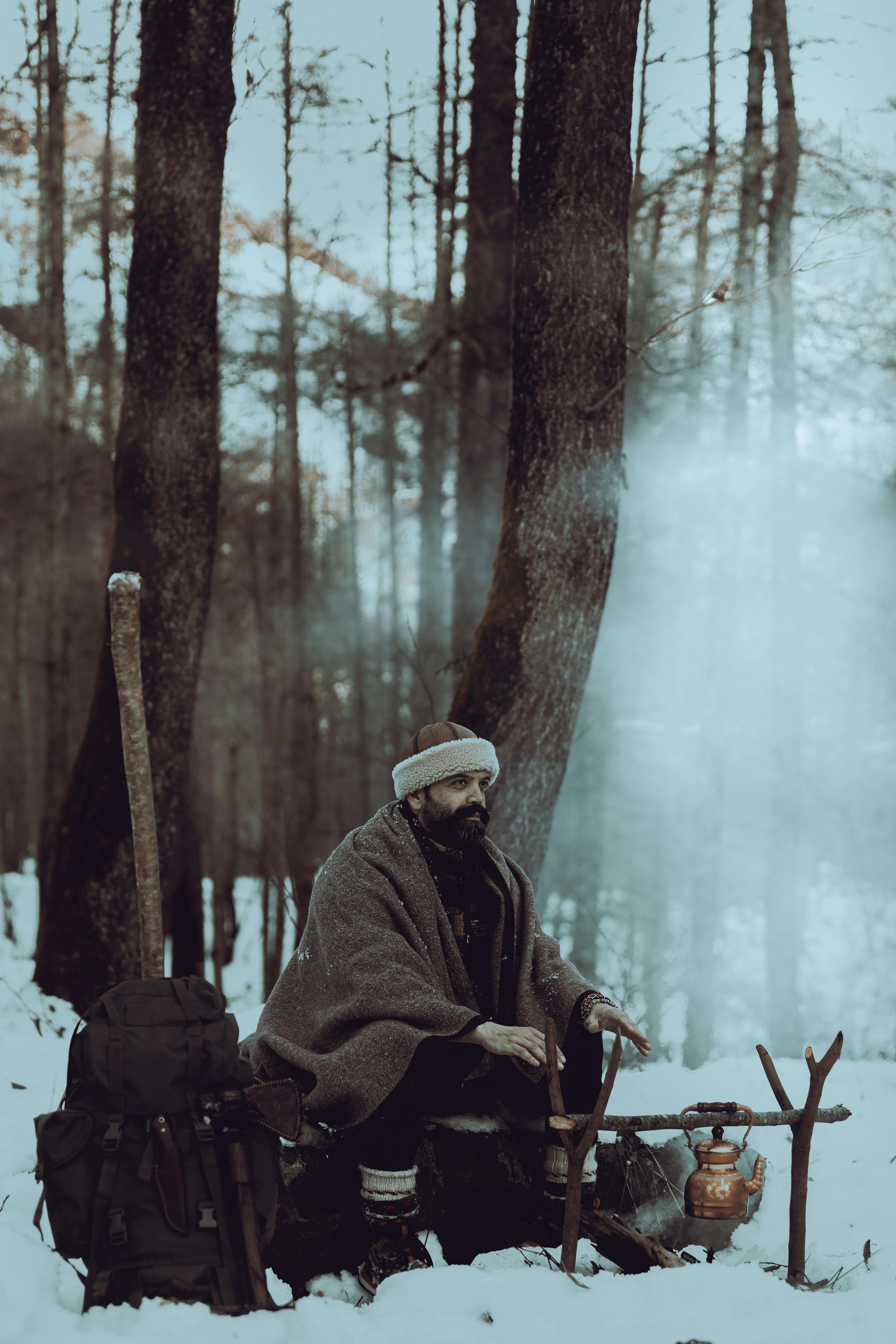 A rugged man in winter clothing rests by a campfire in a snowy forest.