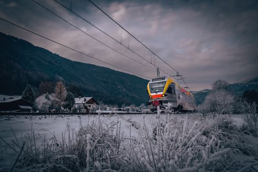 A modern train travels through a scenic snowy mountain landscape under a cloudy sky.