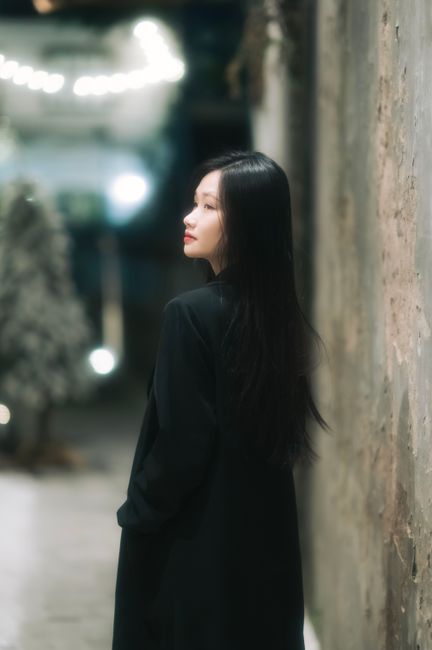 A woman with long hair in an elegant black outfit poses in an urban setting with evening lights.