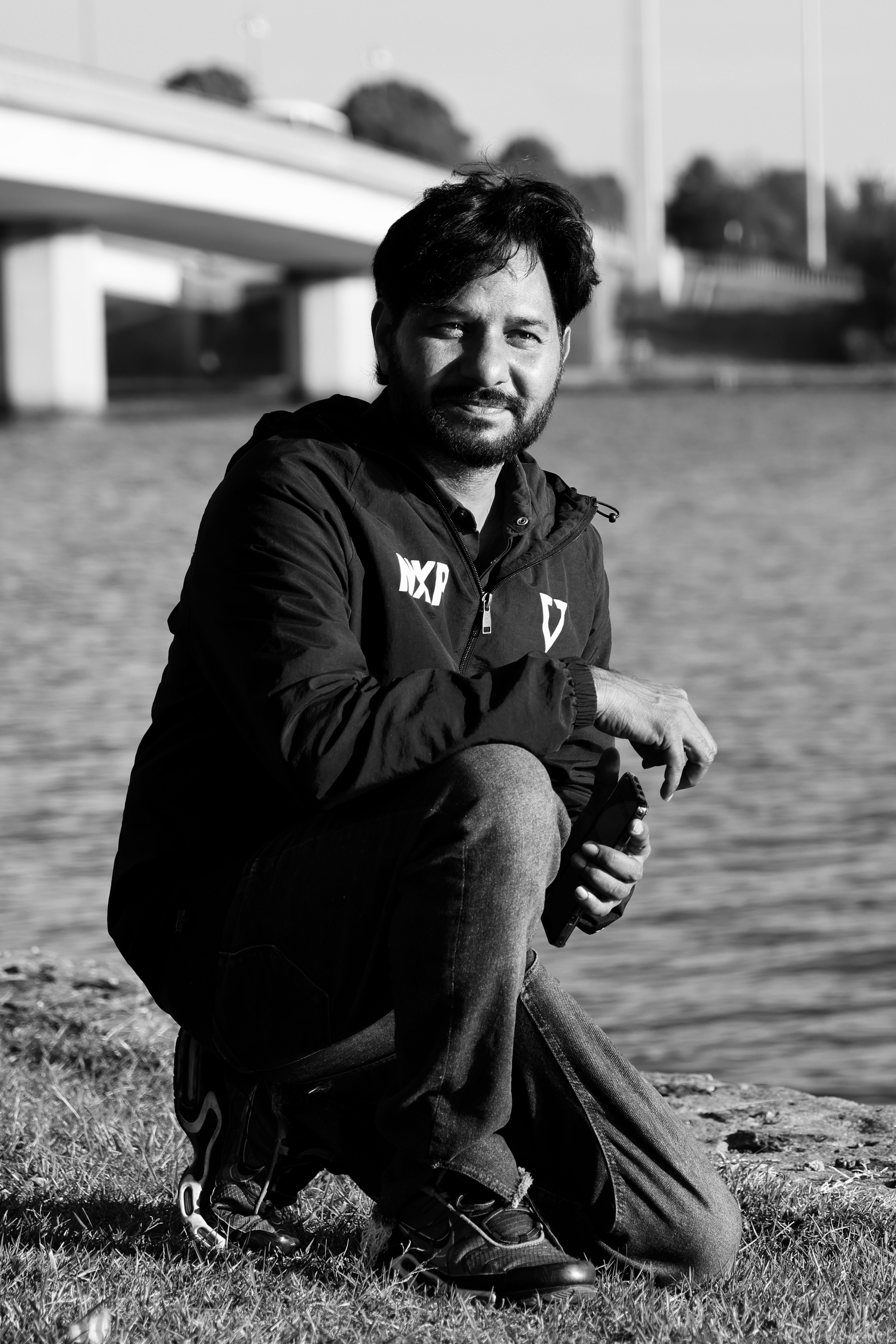 Portrait of a man kneeling by a river with a bridge in black and white. Outdoors setting.