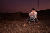 一位时尚男士戴着牛仔帽，坐在沙漠中欣赏日落，展现出时尚与冒险精神