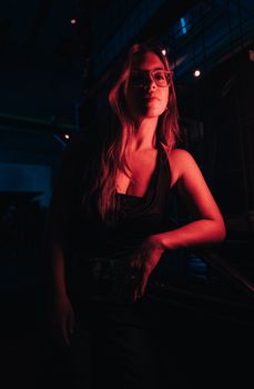 A woman poses stylishly under neon lights in a dark, moody environment.