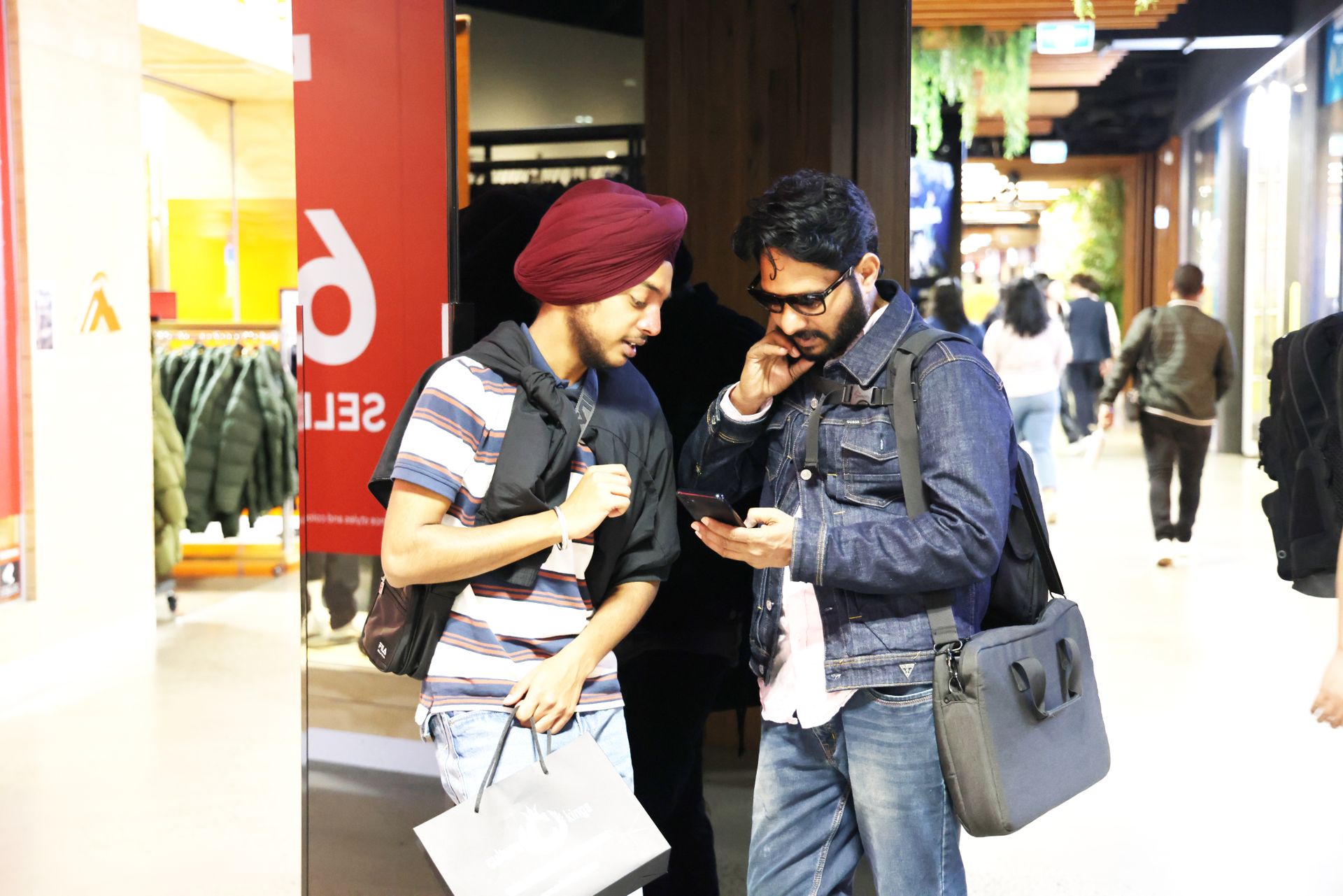 Two men casually shopping and checking a smartphone in a modern mall setting.
