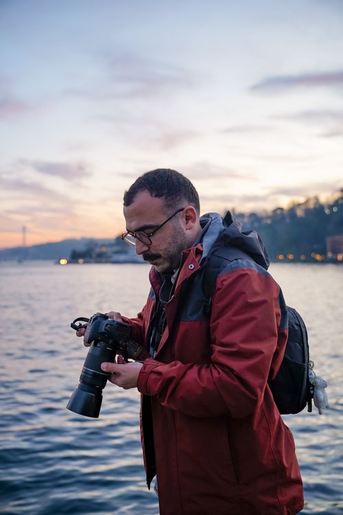 Free Man handling camera near waterfront during twilight, capturing serene landscape. Stock Photo