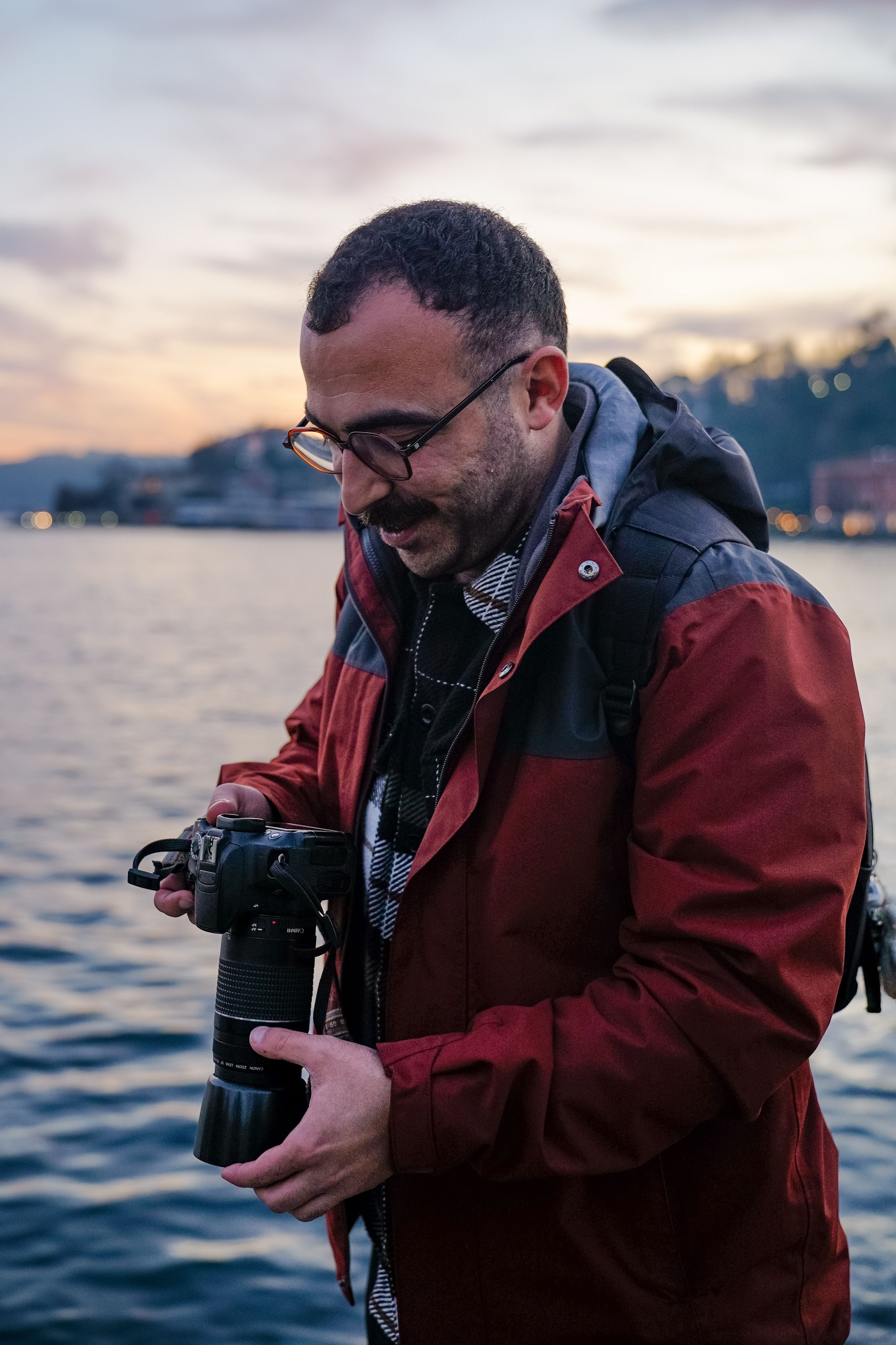 Photographer in red jacket adjusts camera by a lake during twilight.