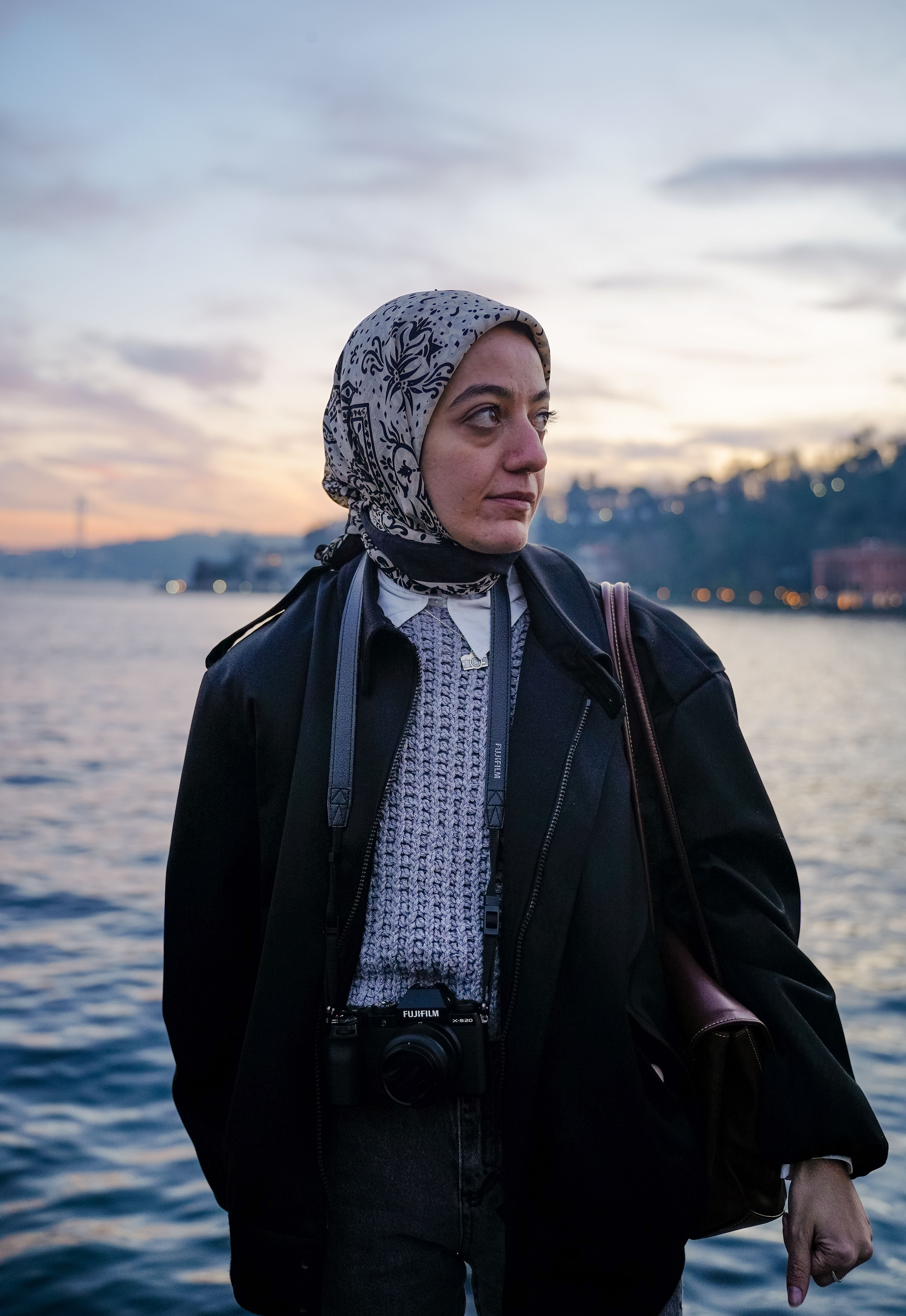 A woman by the water at twilight, holding a camera around her neck, looking contemplative.