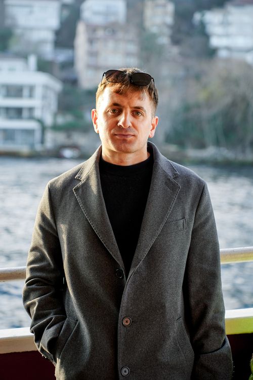 Free A man in a coat poses by a river with buildings in the background. Stock Photo