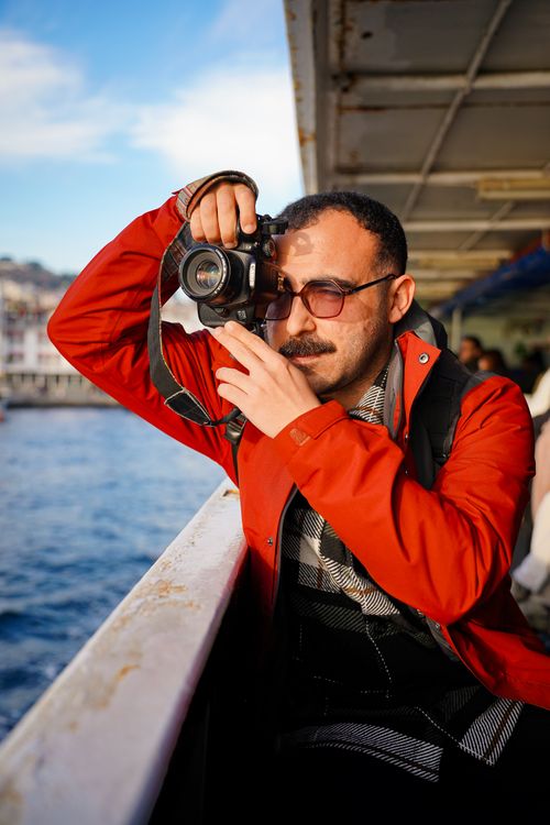 Free Man capturing waterfront views from a ferry with a camera, wearing a red jacket. Stock Photo