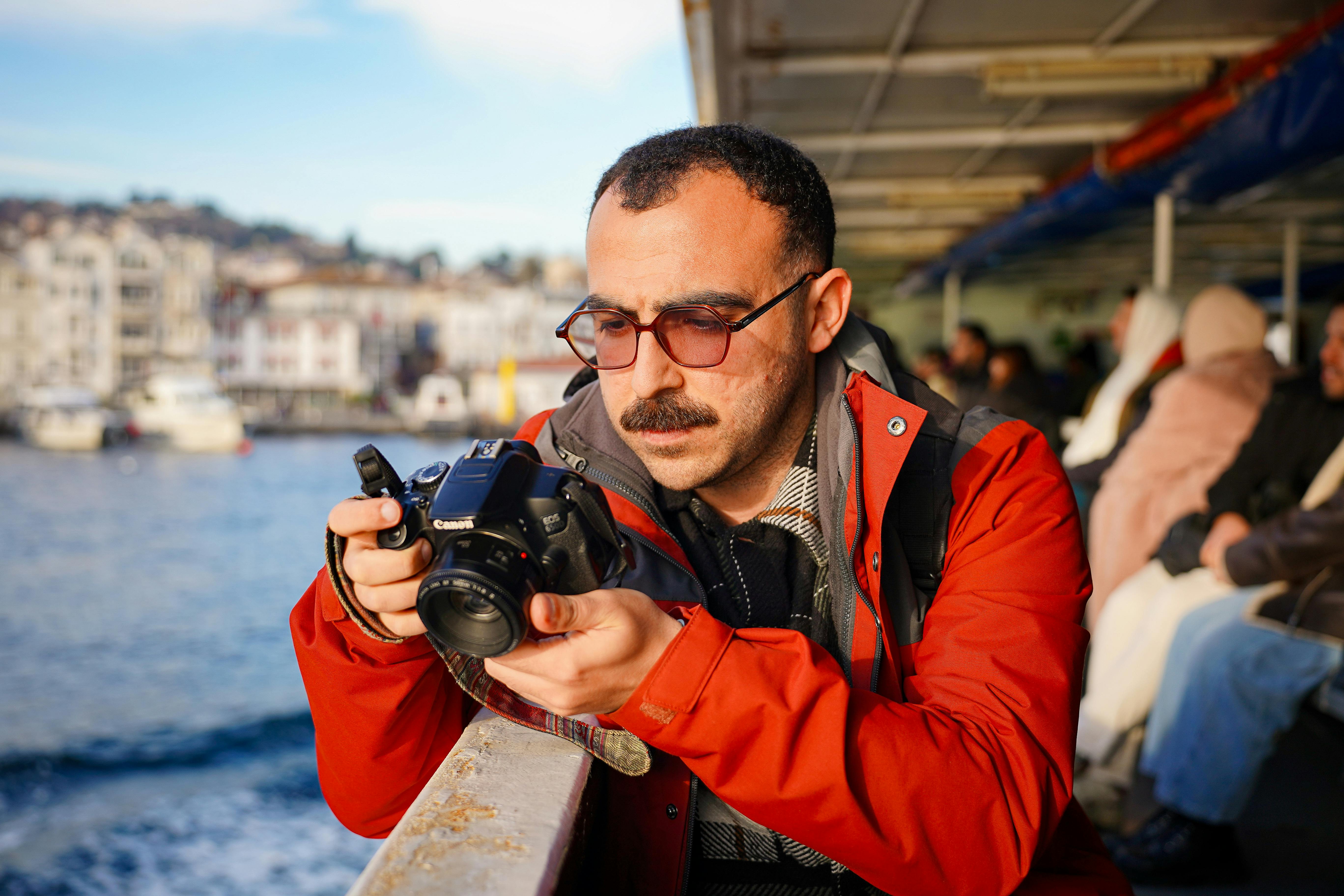 Man in red jacket observing a camera on a ferry with scenic background.