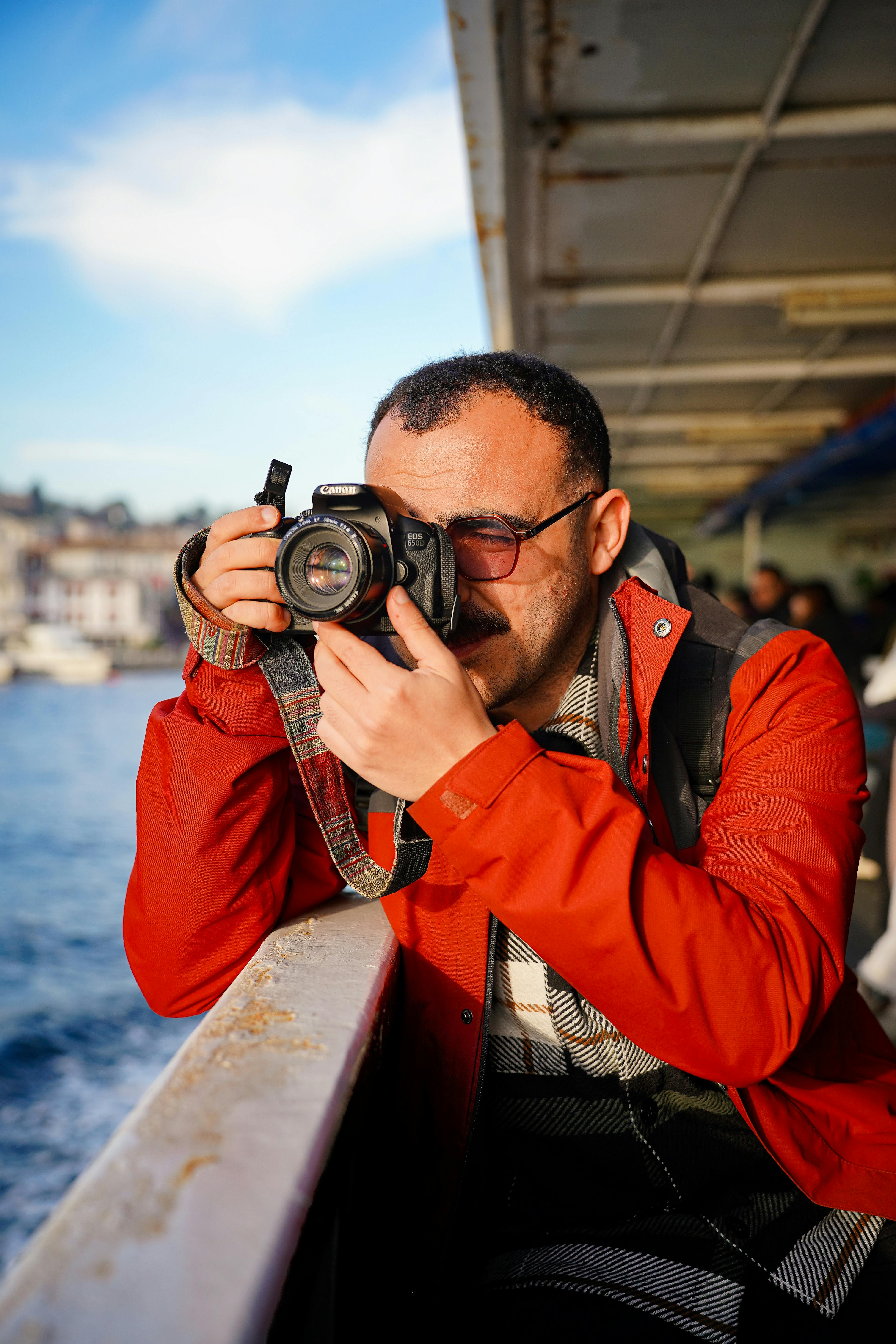 Man in red jacket taking a photo aboard a ferry under clear skies.