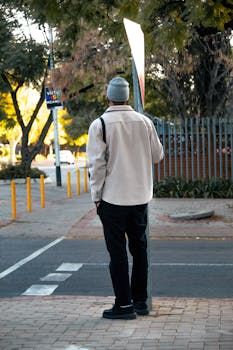 A man in casual attire waits at a crosswalk on a city street during a cool morning.