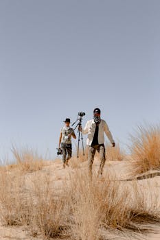 阳光明媚的日子里，两名摄影师带着相机装备徒步穿越沙质沙漠
