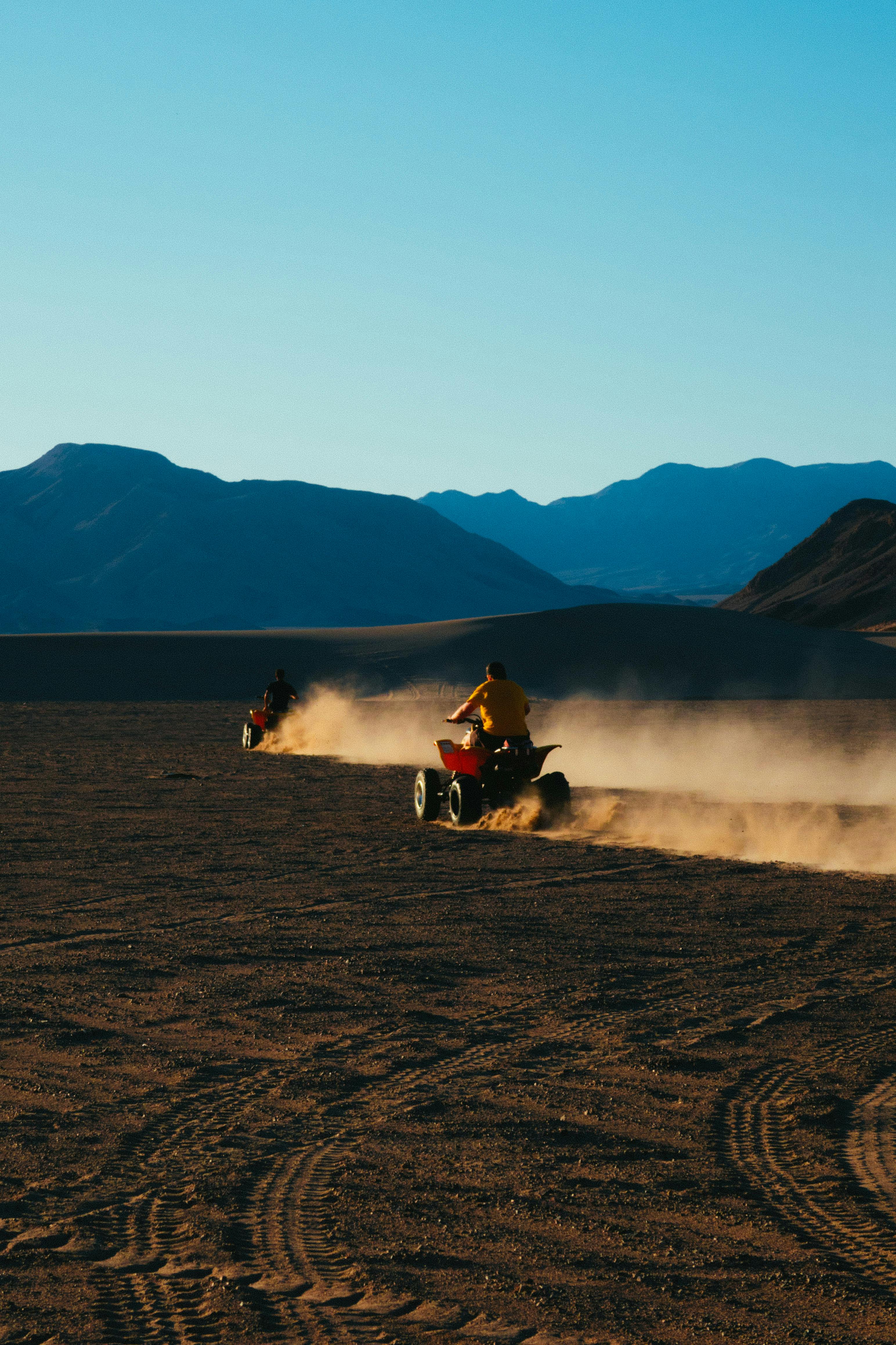 Two riders on ATVs kicking up dust in a vast desert landscape against mountains.