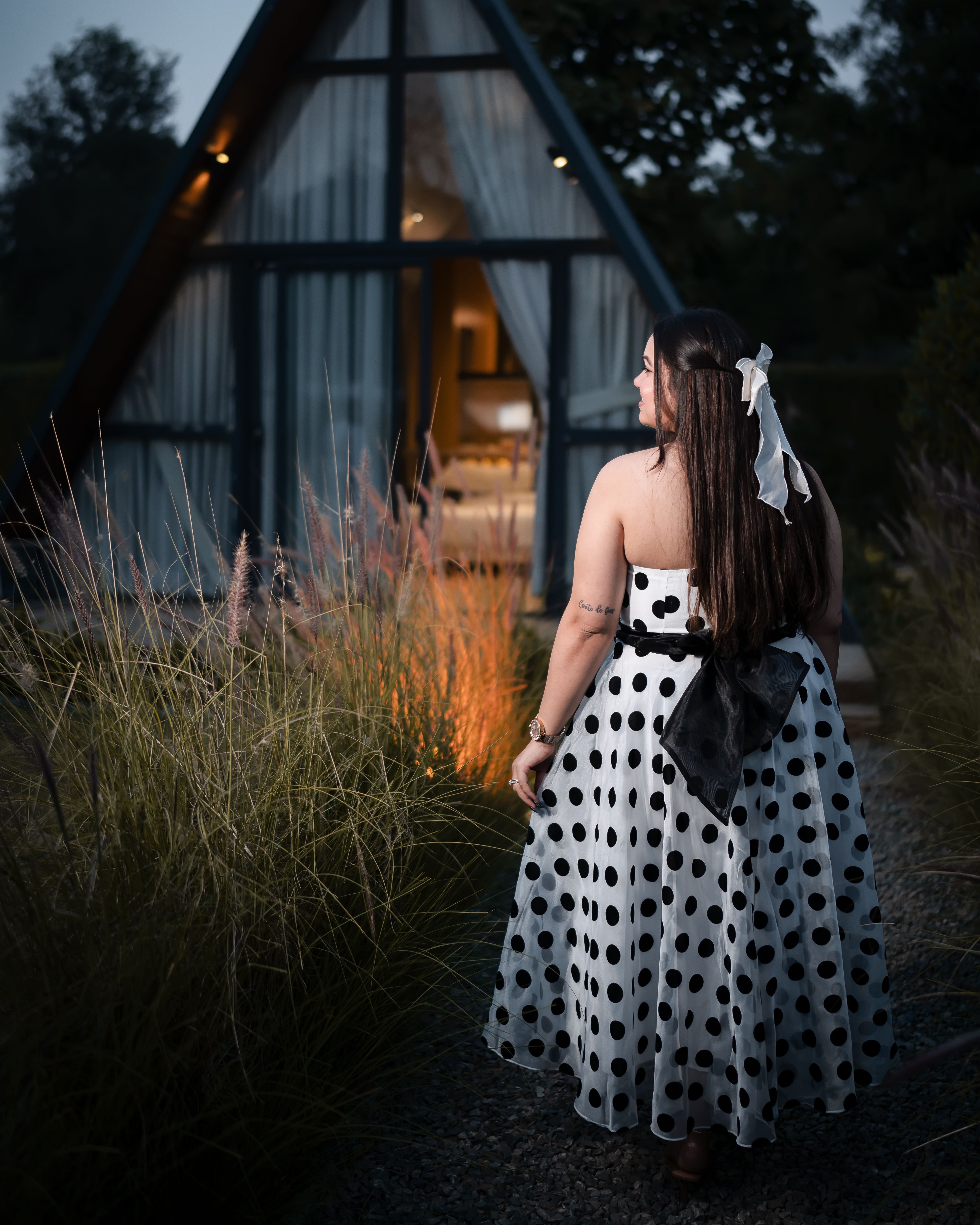 Elegant woman in a polka dot dress stands near a cozy A-frame cabin at dusk, surrounded by tall grass.