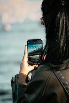 A person photographing a bridge using a smartphone by the water.