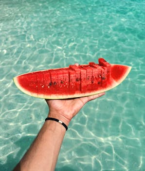 A hand holds a fresh watermelon slice against the backdrop of Thailand's blue waters.